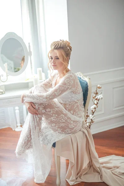 Sabah düğün saç stili ve makyajlı genç ve güzel bir gelin yatak odası masasının yanında, yere kadar uzanan beyaz dantelli bir ceketle. düğün sabahı