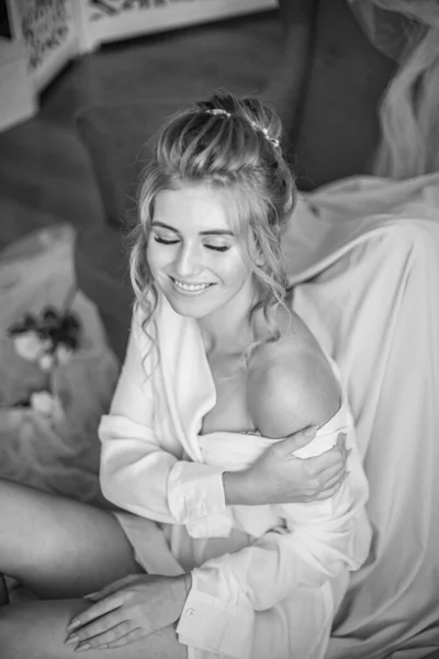 Young beautiful bride with wedding hairdo and makeup in the morning wearing a white silk negligee in the bedroom. wedding morning