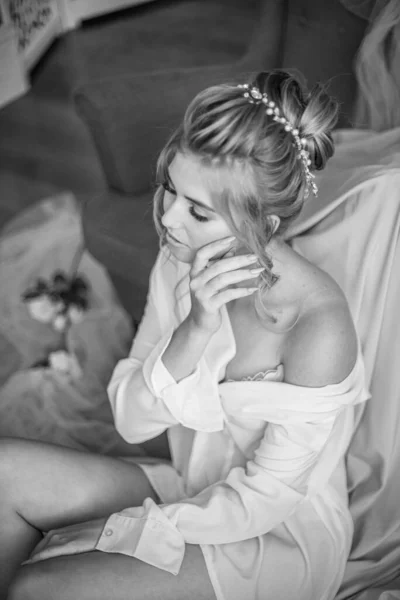 Young beautiful bride with wedding hairdo and makeup in the morning wearing a white silk negligee in the bedroom. wedding morning