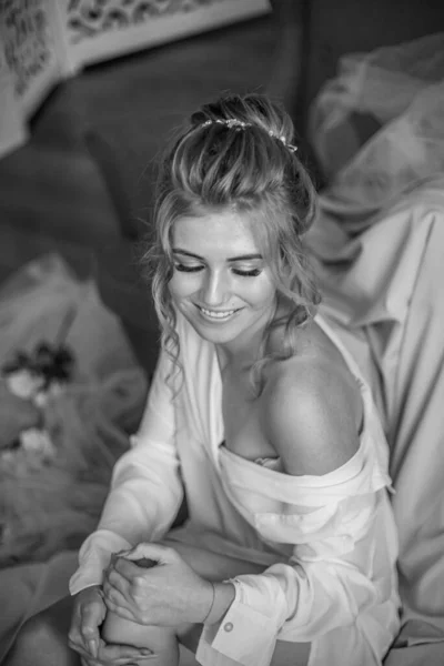 Young beautiful bride with wedding hairdo and makeup in the morning wearing a white silk negligee in the bedroom. wedding morning