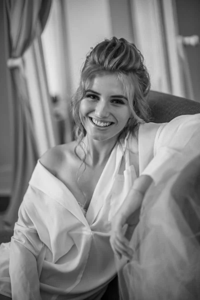 Young beautiful bride with wedding hairdo and makeup in the morning wearing a white silk negligee in the bedroom. wedding morning