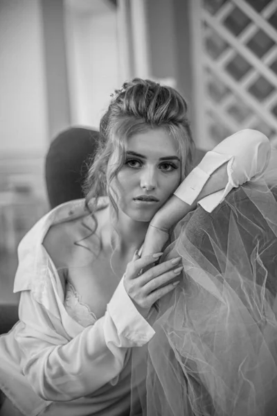 Young beautiful bride with wedding hairdo and makeup in the morning wearing a white silk negligee in the bedroom. wedding morning