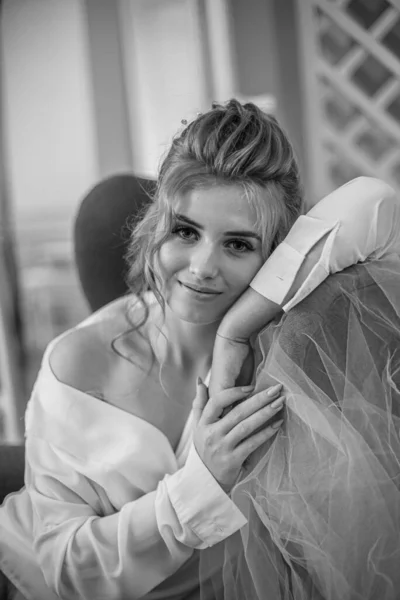 Young beautiful bride with wedding hairdo and makeup in the morning wearing a white silk negligee in the bedroom. wedding morning