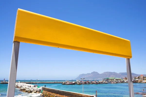 Yellow train station sign against a blue sky with the Kalk Bay harbour