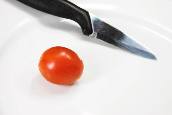 Uma faca e um tomate cereja em um prato branco . — Fotografia de Stock