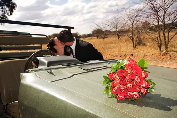 Red bouquet of roses on the bonnet of a car, just married bridal couple kissing in the background — Stock Photo, Image