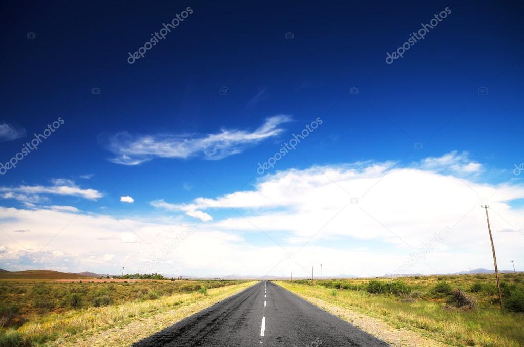 Long straight road under a dramatic dark blue saturated sky with some fine white clouds, and lined with some greenery and small shrubs