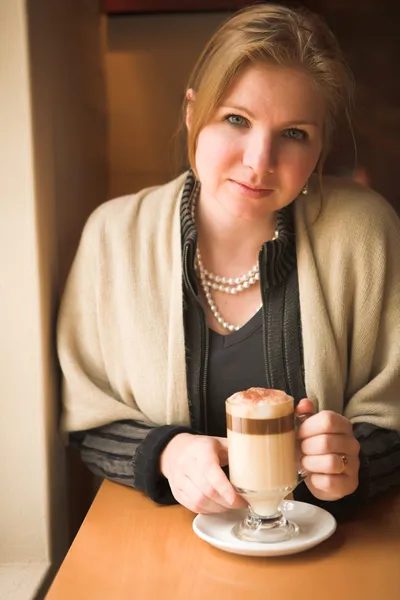 Rubia caucásica mujer adulta sentada junto a una ventana de luz natural en el comienzo del invierno bebiendo café con leche caliente — Foto de Stock