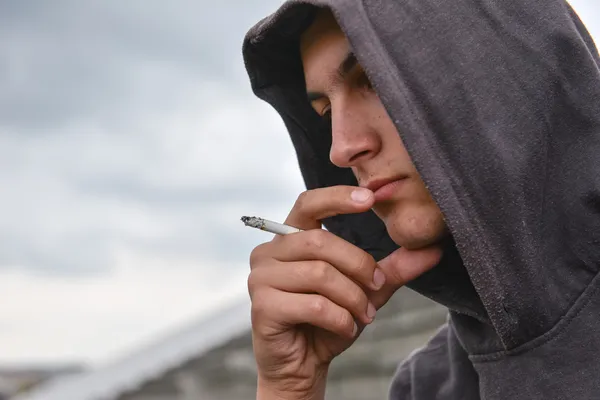 Promyšlený a ustaraný dospívající chlapec s černým hoodie je kouření cig — Stock fotografie