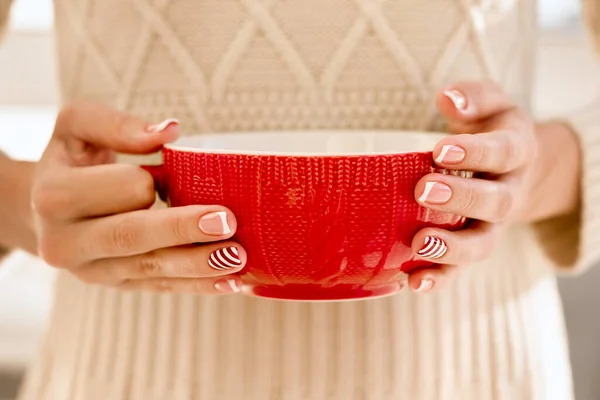 Womans hands with french manicure and candy cane pattern on the nails. Woman with beautiful manicure holding big red knitted cup. The concept of cozy Christmas holidays and New Year. — 图库照片