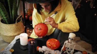 Cadı şapkalı bir kadın turuncu balkabağına korkunç bir yüz çiziyor. Jack O Lantern. Cadılar Bayramı gecesini kutluyoruz.