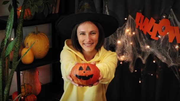 Счастливого Хэллоуина. Улыбающаяся молодая ведьма держит тыкву с раскрашенным страшным лицом — стоковое видео