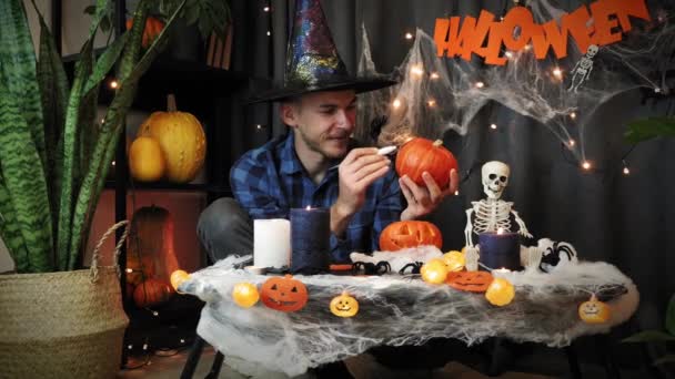 Hombre gracioso en sombrero de bruja dibujando cara de miedo en calabaza. Saludos de Halloween — Vídeos de Stock