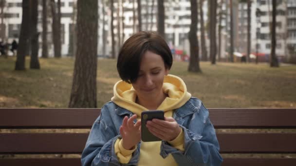 Усміхнена молода жінка використовує смартфон, спілкується онлайн, прокручує новини в Інтернеті в парку — стокове відео
