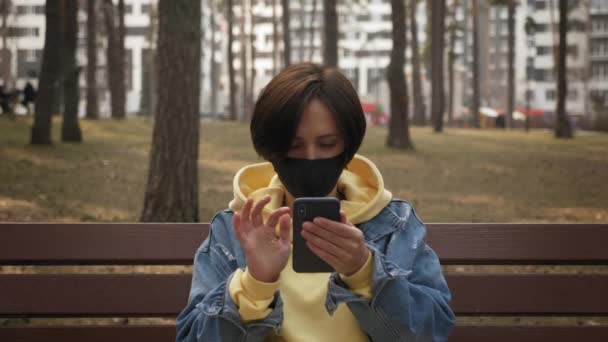 Mujer joven con mascarilla protectora está sentado en el banco en el parque y la pantalla del teléfono inteligente de desplazamiento — Vídeo de stock