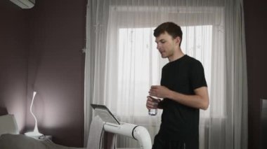 Adam koşu bandında yürürken su içiyor. Sporcu evde egzersiz yapıyor.