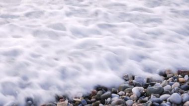 Kumsalda çakıl taşları, yakın çekim. Deniz dalgaları çakıl taşları plajına ulaşıyor, beyaz köpük oluşturuyor.