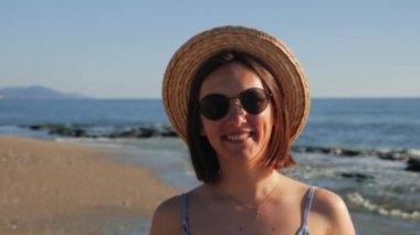 Hasır şapkalı mutlu kız kumsalda güneşlenip esinlenerek kameraya bakıyor.
