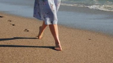 Güneşli yaz gününde kumlu sahilde çıplak ayakla yürüyen, el sallayan elbiseli bir kadın ayak izleri bırakıyor.