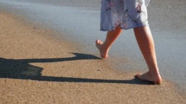 Gün batımında kumsalda yürüyen bir kadın. İnce kadın bacakları ve ayakları deniz sahilinde yürüyor.