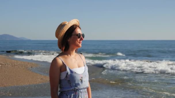 Очаровательная девушка в шляпе и солнцезащитных очках стоит на берегу моря и смотрит на горизонт — стоковое видео