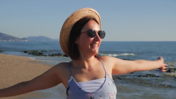 Очаровательная девушка в соломенной шляпе наслаждается океанским бризом на пляже. Счастливая улыбающаяся женщина на пляже — стоковое видео