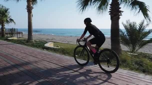 Cyclist rides road bicycle through palm trees on promenade of sandy beach. Woman cycling bike at sunny summer day — Stock Video