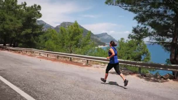 Woman jogging across a highway overpass in mountains with sea on background — стоковое видео