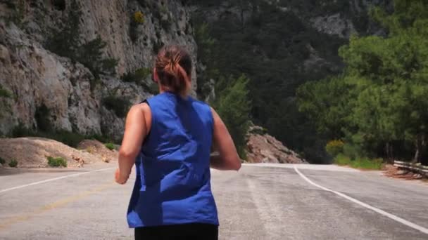 Volver seguir disparo de mujer joven corriendo por carretera rodeado de verdes montañas — Vídeo de stock