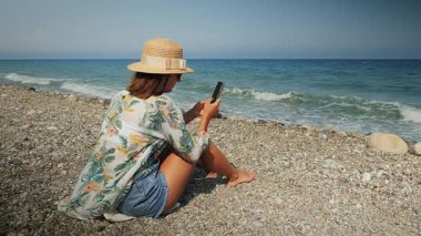 Hasır şapkalı çekici bir kadın kumsalda oturuyor, rahatlıyor ve akıllı telefon kullanıyor.