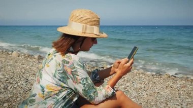 Genç bir bayan sahilde, rahatlıyor ve akıllı telefon kullanıyor. Yaz tatili