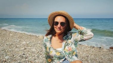 Hasır şapkalı, rüzgarda dalgalanan saçlarıyla gülümseyen kız güneşli bir günde kumsalda oturuyor.