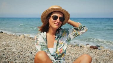 Hasır şapkalı, dalgalı saçlı ve plajda oturan tropikal yaz gömlekli bir kadın.