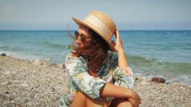 Yazlık şapkalı mutlu kadın rüzgarlı bir günde kumsalda oturmuş güneşli plaj gününün tadını çıkarıyor.