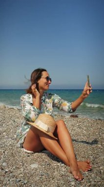 Hasır şapkalı, kumsalda saçlarını sallayan, rüzgarlı bir günde selfie çeken mutlu bir kadın.