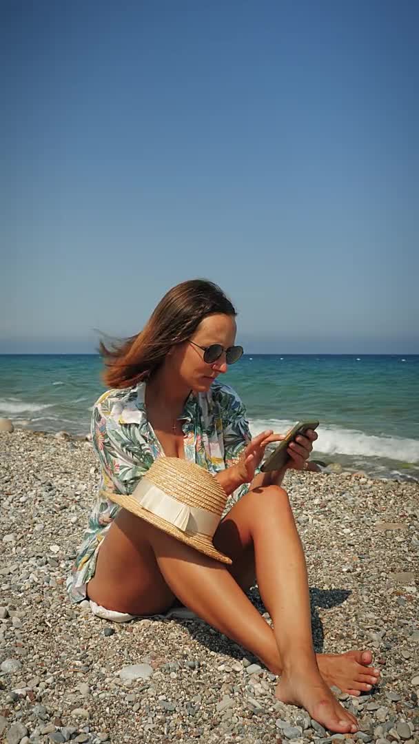 Gelooide jonge vrouw zitten op het strand en met behulp van smartphone, verticale video — Stockvideo
