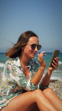 Kaygısız hippi kız sahilde oturuyor ve akıllı telefon kullanıyor.