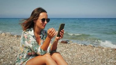 Kumsalda oturan, akıllı telefon kullanan, tropikal tatilin tadını çıkaran hoş bir kız.