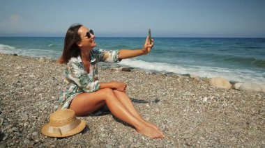 Mutlu beyaz kadın sahilde oturuyor ve akıllı telefondan selfie çekiyor.