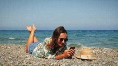 Neşeli esmer kız rüzgarlı bir yaz gününde kumsalda uzanıyor ve akıllı telefon kullanıyor.