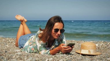 Sevimli hippi kız sahilde uzanıyor, akıllı telefon kullanıyor ve internetten iletişim kuruyor.