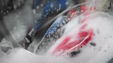 Çamaşır makinesi, kapat. Beyaz sabun köpükleriyle çamaşır makinesinde dönen renkli çamaşırlar. Evde çamaşır yıkayan bir kadın.