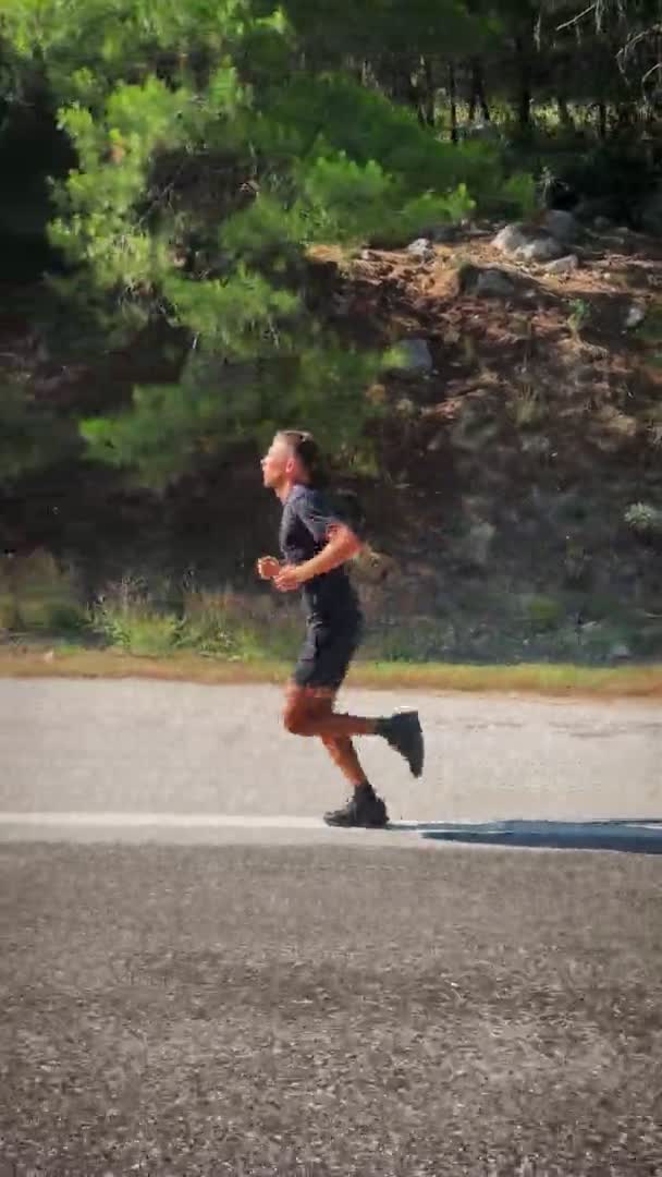 屋外で走ってる男 日当たりの良い夏の日に山の森を介してアスファルトの道路上に筋肉のジョギングに適合する若い男性アスリート 山の中で黒のランニングウェアのトレーニングでスポーツマン — ストック動画
