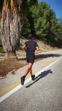 Dışarıda koşan bir sporcu. Dikey video. Siyah spor giysili bir adam yolda koşuyor. Genç formda bir erkek triatlet dağlarda koşuya hazırlanıyor. Spor konsepti. Fitness egzersizi