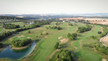 Golf sahasının havadan görünüşü. Golf sopası üzerinde uçan bir dron. Akşam gün batımında yeşil golf sahaları. Eğlence etkinliği. Spor konsepti. Tepelerde yeşil ağaçlar ve göl olan güzel doğa manzarası