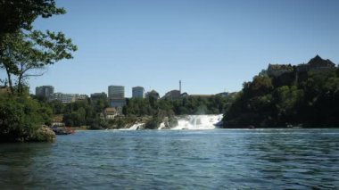 Waterfall of Rhine river with massive water power and ships with passengers and tourists, timelapse. Rhine falls and surrounding nature landscape. Rheinfall waterfall on Rhine river in Switzerland