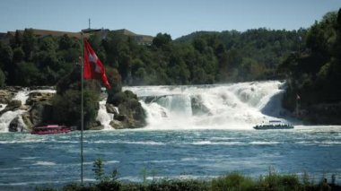 Massive waterfall on Rhine river with waving flag of Switzerland. Flag of Switzerland fluttering in wind against Rhine Falls with power water stream. Amazing nature landscape. Tourism and travel