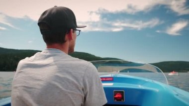 Man driving electric boat on lake. Young male in black hat turning wheel on yacht, floating along beautiful mountain lake. Summer vacation concept. Sailing and travelling concept. Holiday weekend