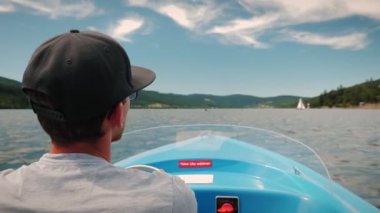 Back view of man steering in yacht on sunny day. Man driving electric boat along lake surrounded by green trees and mountains. Male driving river vessel, relaxing and resting on weekend