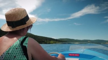 Dreamy woman sailing boat on lake at sunny day during summer vacation. Cheerful charming woman in straw hat floating on electric tourist ship along mountain lake with green trees and hills on horizon